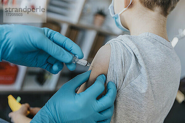 Arzt mit Schutzhandschuh bei der Injektion des Impfstoffs COVID-19 in den Arm eines Jungen in einer Klinik