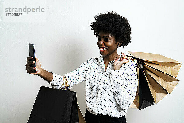 Lächelnde junge Frau mit Einkaufstaschen  die ein Selfie macht  während sie vor einem weißen Hintergrund steht