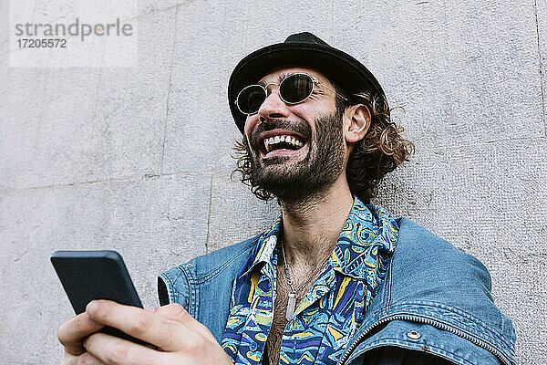 Lachender gut aussehender Mann mit Sonnenbrille  der sein Smartphone vor einer Wand hält