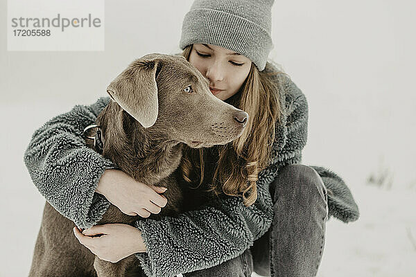 Porträt eines Mädchens im Teenageralter  das im Schnee hockt und einen Labrador Retriever umarmt