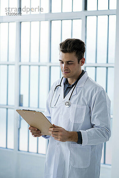 Männlicher Arzt betrachtet Dokumente und hält ein Klemmbrett im Krankenhaus