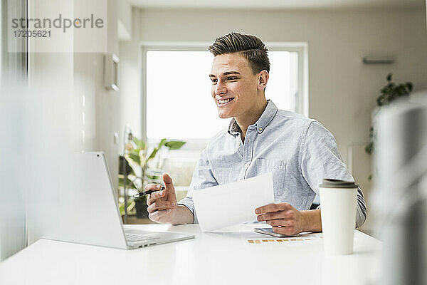 Glücklicher Geschäftsmann mit Dokument  der einen Videogespräch über einen am Schreibtisch sitzenden Laptop führt