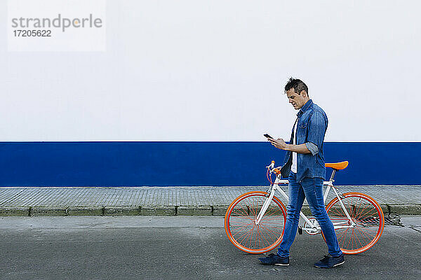 Älterer Mann  der ein Mobiltelefon benutzt  während er auf der Straße mit dem Fahrrad an der Wand spazieren geht