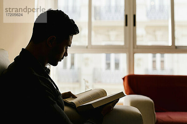 Junger Mann sitzt auf dem Sofa und liest ein Buch zu Hause