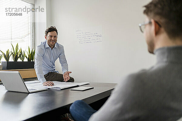 Glücklicher Geschäftsmann  der mit einem Kollegen am Schreibtisch im Büro über einen Bauplan diskutiert