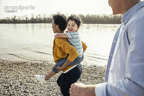 Fröhlicher Junge genießt es  von seiner Mutter huckepack genommen zu werden  während sein Vater am Seeufer spazieren geht