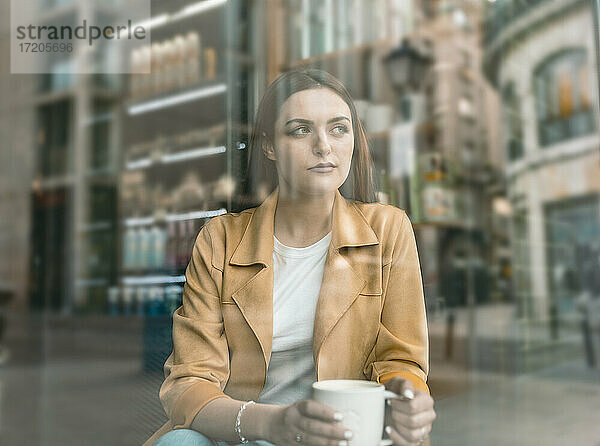 Nachdenkliche junge Frau  die in einem Café sitzt  gesehen durch ein Glasfenster