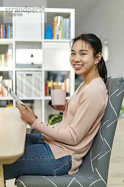 Junge Frau mit digitalem Tablet lächelnd und hält Kaffeetasse sitzen zu Hause