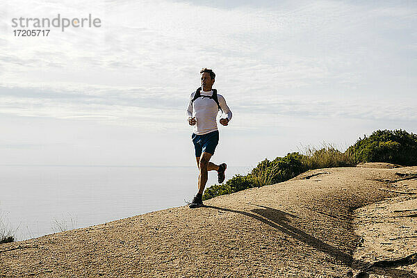 Sportlicher Mann läuft auf unbefestigtem Weg gegen bewölkten Himmel an einem sonnigen Tag