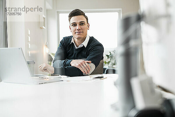 Lächelnder junger Geschäftsmann sitzt am Schreibtisch im Home Office