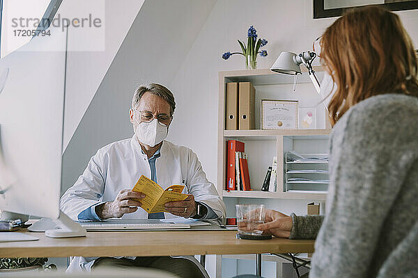 Arzt mit Schutzmaske prüft im Büro sitzend den Impfpass eines Patienten