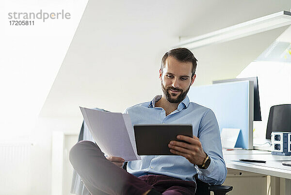 Lächelnder Unternehmer mit digitalem Tablet und Papier im Büro sitzend
