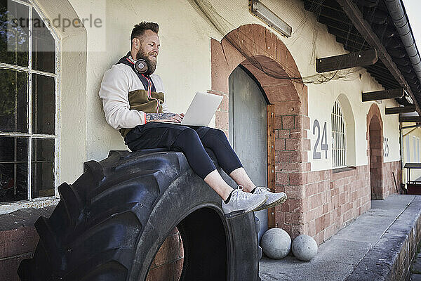 Männlicher Fitnesstrainer  der einen Laptop benutzt  während er auf einem Reifen vor einem Fitnessstudio sitzt