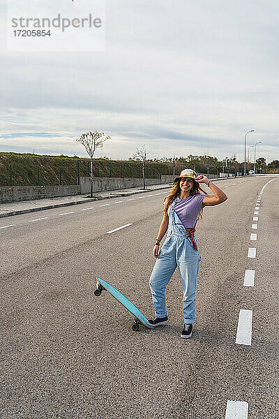 Lächelnde Frau mit Hut steht mit Skateboard auf der Straße