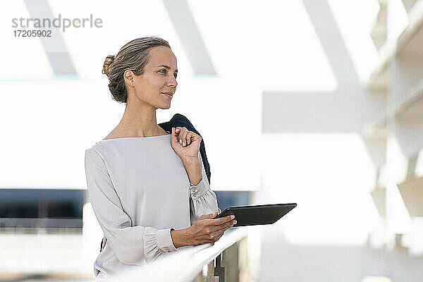 Geschäftsfrau mit digitalem Tablet und Jacke schaut weg  während sie auf einer Büroterrasse steht