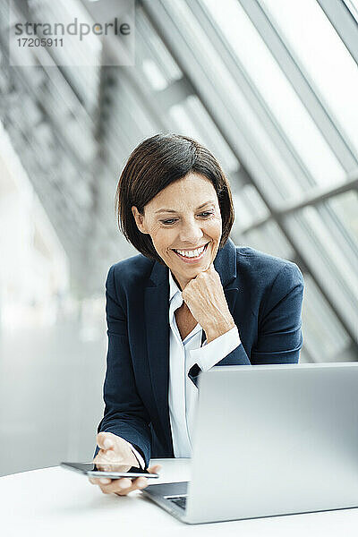 Lächelnde Geschäftsfrau mit Hand am Kinn  die im Korridor am Laptop arbeitet