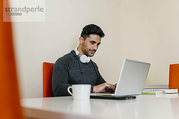 Junger Mann mit Kopfhörer  der einen Laptop benutzt  während er am Tisch im Wohnzimmer sitzt