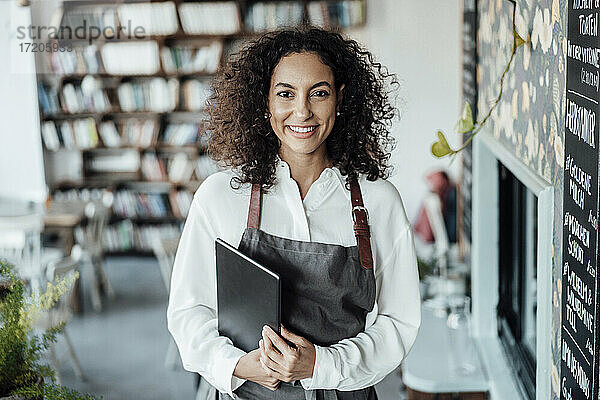 Lächelnde Frau mit Schürze  die ein Buch hält  während sie in einem Café steht