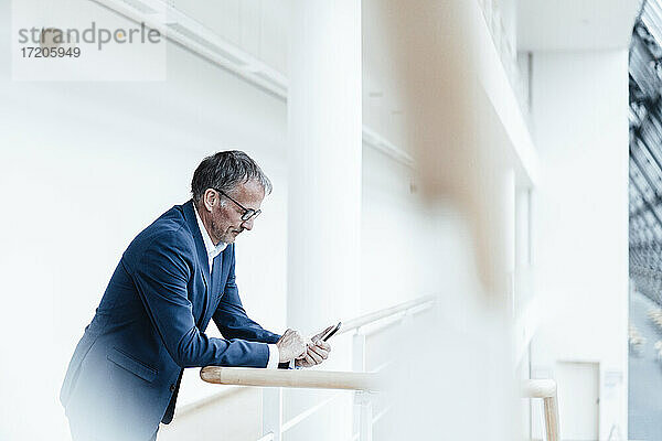 Älterer männlicher Geschäftsmann  der sein Smartphone benutzt  während er sich auf das Geländer im Büroflur stützt