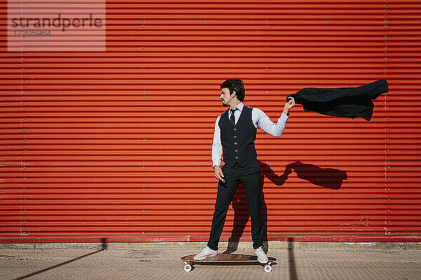 Männlicher Unternehmer hält Blazerjacke  während er auf einem Longboard an der Wand steht