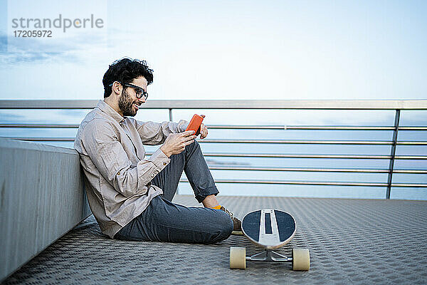 Lächelnder Geschäftsmann  der ein Mobiltelefon auf einem Longboard benutzt  während er auf einem Pier sitzt und sich gegen eine Bank lehnt