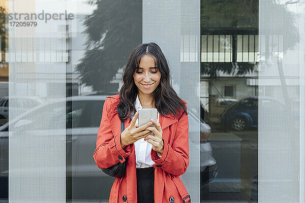 Lächelnde Frau in rotem Trenchcoat benutzt ein Smartphone  während sie an einer Glaswand steht