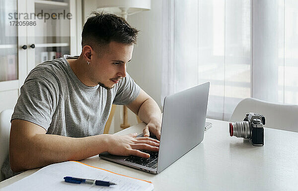 Junger männlicher Berufstätiger  der einen Laptop benutzt  während er ein Video mit einer Digitalkamera im Heimbüro aufnimmt