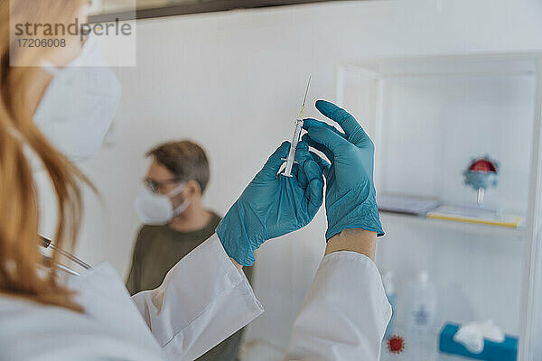 Allgemeinmediziner bei der Vorbereitung des COVID-19-Impfstoffs für den im Hintergrund sitzenden Patienten im Untersuchungsraum