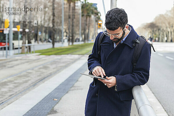 Männlicher Geschäftsmann  der ein digitales Tablet benutzt und sich am Geländer eines Bahnhofs anlehnt