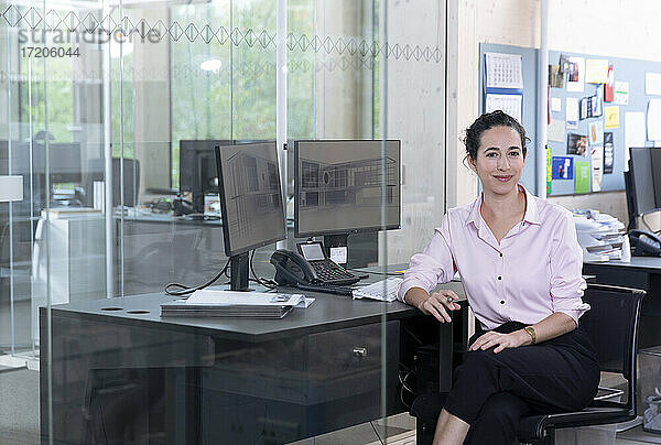 Lächelnde Geschäftsfrau sitzt auf einem Stuhl am Schreibtisch im Büro