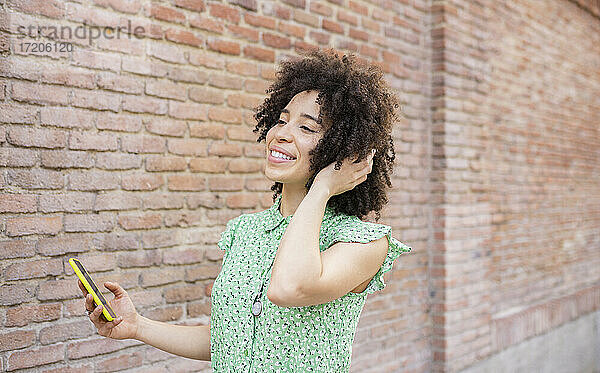 Lächelnde Frau mit Hand in den Haaren  die ihr Smartphone an einer Backsteinmauer hält