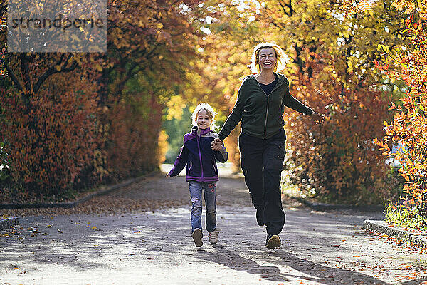 Glückliche Mutter und Tochter halten sich an den Händen und laufen im Herbst im Park