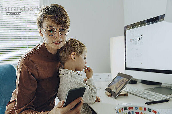 Eine berufstätige Mutter benutzt ein Smartphone  während ihr Sohn ein Video auf einem digitalen Tablet ansieht
