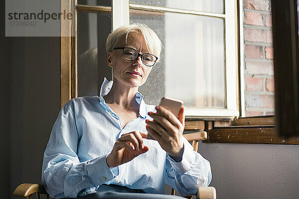 Reife Geschäftsfrau  die ein Mobiltelefon benutzt  während sie auf einem Stuhl am Fenster im Heimbüro sitzt