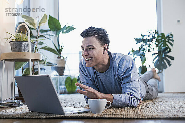 Lächelnder junger Mann  der auf einem Teppich liegt und bei einem Videoanruf über einen Laptop gestikuliert