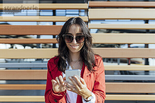 Lächelnde weibliche Fachkraft mit Sonnenbrille schreibt eine SMS  während sie an einer Holzwand steht