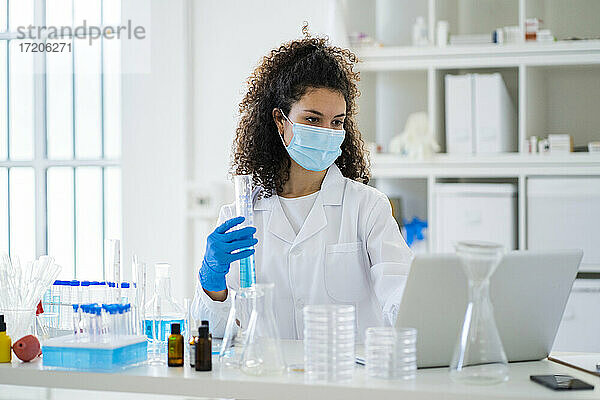 Junge Forscherin mit Reagenzglas bei der Arbeit am Laptop in einem Krankenhaus während einer Pandemie