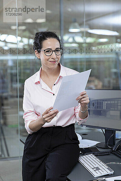 Lächelnde Geschäftsfrau mit Strategie sitzt am Schreibtisch im Büro