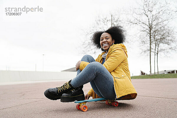 Lächelnde Frau auf dem Skateboard sitzend auf dem Fußweg gegen den klaren Himmel
