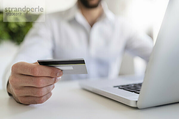Unternehmer mit Laptop  der eine Kreditkarte hält  während er am Schreibtisch im Büro sitzt