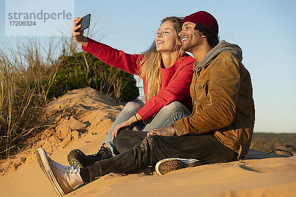 Lächelndes Paar  das ein Selfie mit dem Handy macht  während es auf einer Sanddüne sitzt