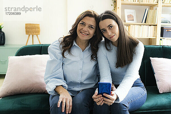 Lächelnde Tochter mit Smartphone bei der Mutter auf dem Sofa zu Hause