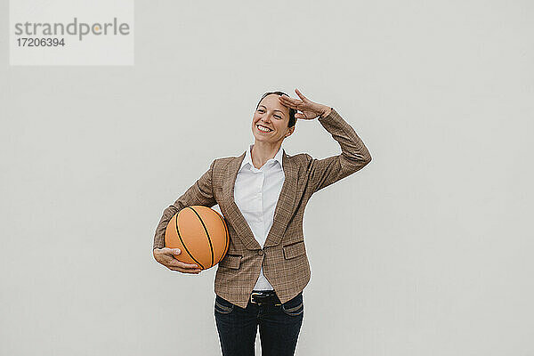 Lächelnde weibliche Fachkraft mit Basketball  die vor einer weißen Wand steht und grüßt