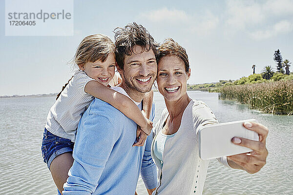 Lächelnde Frau nimmt Selfie mit Familie am See an einem sonnigen Tag