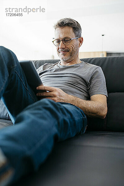Lächelnder reifer Mann  der ein digitales Tablet benutzt  während er auf dem Sofa sitzt