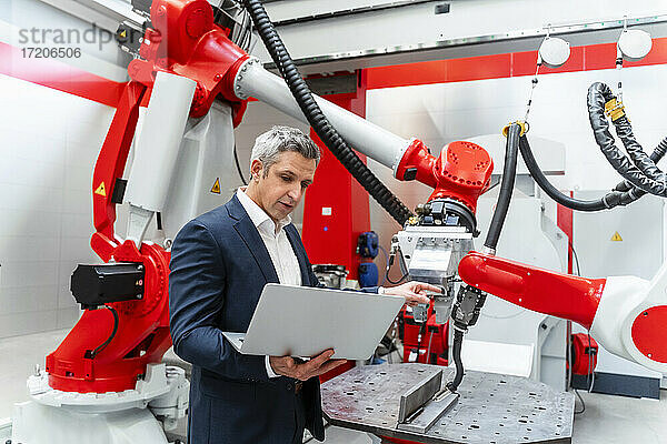 Ein männlicher Ingenieur zeigt auf einen Roboter  während er eine Videokonferenz über einen Laptop in einer Fabrik abhält.