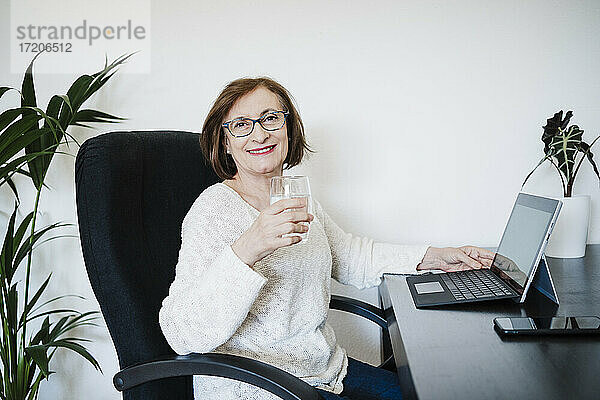 Lächelnde ältere Frau mit Laptop  die Wasser trinkt  während sie auf einem Stuhl zu Hause sitzt