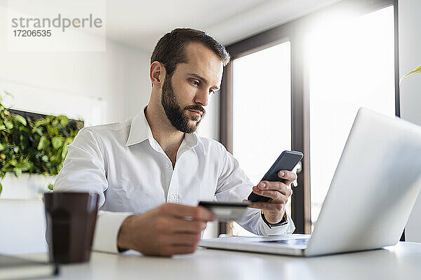 Berufstätiger mit Laptop und Kreditkarte bei der Nutzung eines Mobiltelefons im Büro