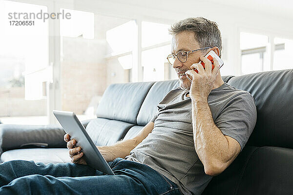 Älterer Mann  der auf dem Sofa im Wohnzimmer mit einem Mobiltelefon spricht und ein digitales Tablet benutzt