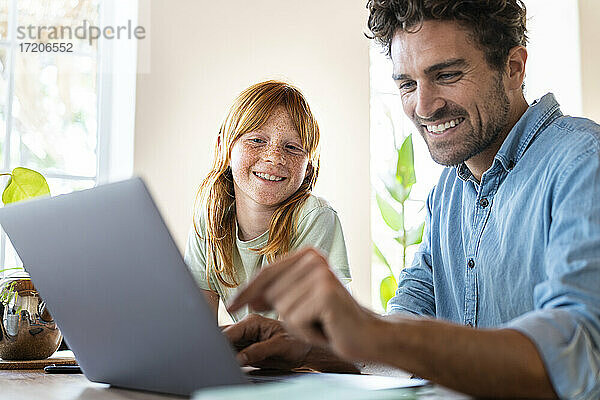 Lächelndes rothaariges Mädchen mit Vater schaut im Wohnzimmer auf einen Laptop
